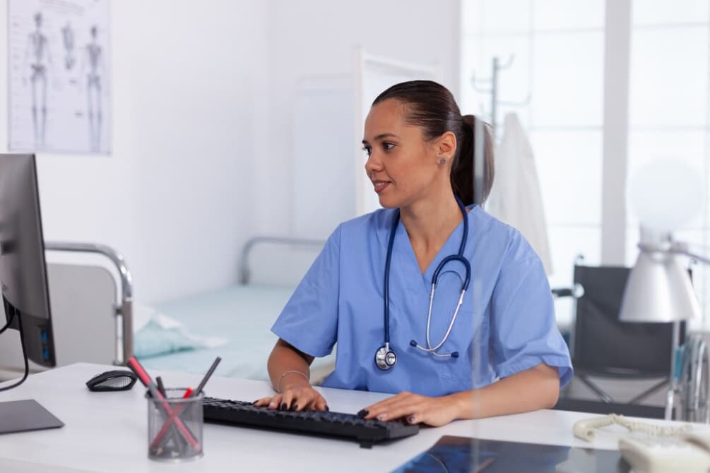 A nurse sits at a desk in a clinic, working on a computer