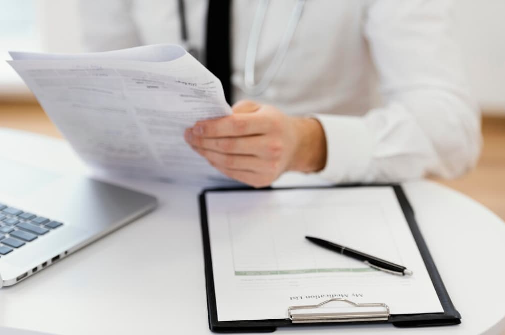 Close-up of a doctor reviewing documents with a laptop beside them