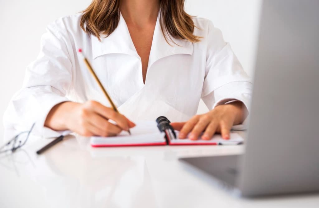 A person in a white coat is writing notes beside a laptop