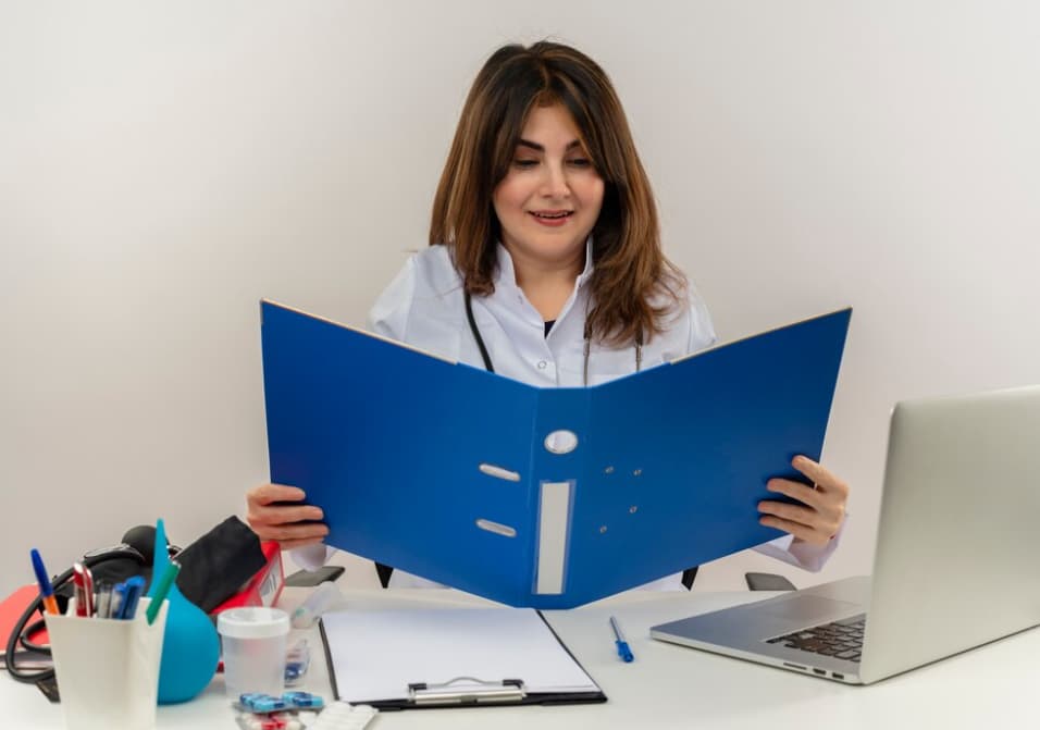 A smiling female doctor reading a file at her desk with medical supplies