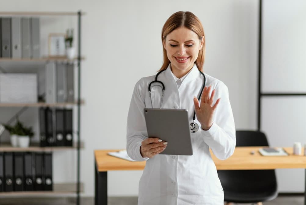 A smiling female doctor waving while holding a tablet